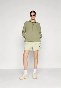 Pulover verde din fleece cu fermoar pe jumătate și buzunar tip cangur, asortat cu pantaloni scurți cargo bej deschis, având buzunare frontale și croială lejeră.