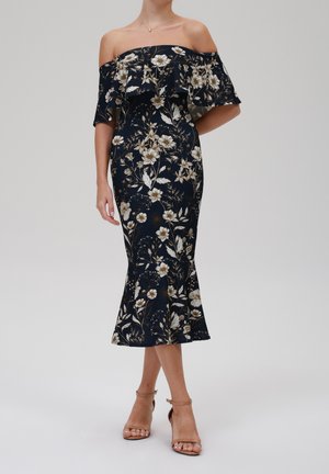 Frau trägt ein marineblaues Midikleid mit Carmenausschnitt, weißem und beigem Blumenmuster sowie nudefarbene, knöchelhohe Sandalen mit Absatz und Riemchen.