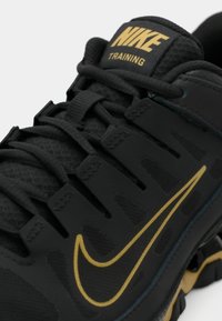 Černá tréninková bota Nike s texturovaným syntetickým svrškem, zlatým logem a černými tkaničkami. Obsahuje zesílené očka a viditelnou vzduchovou jednotku v podrážce.