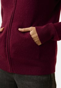 Burgundowa wełniana kardigan z prążkowanymi mankietami i dołem, z dwoma przednimi kieszeniami i zamkiem błyskawicznym na całej długości. Gładka struktura.