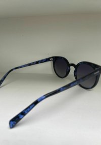 Lunettes de soleil rondes avec un motif marbré bleu, verres foncés et montures en plastique résistant. La branche droite arbore le logo de la marque.