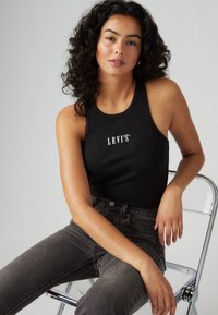 Črna rebrasta majica s tankimi naramnicami z logotipom "LEVI'S®" v beli barvi, v kombinaciji s temno sivo jeansom. Model pozira sedeč na prozorni stoli.