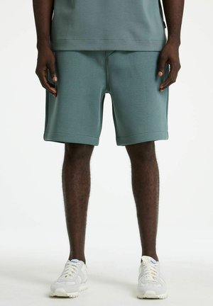 Ein Mann trägt türkisfarbene Shorts, ein passendes türkisfarbenes Hemd und weiße Turnschuhe und steht vor einem einfarbigen weißen Hintergrund.