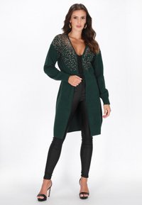 Langer grüner Cardigan mit tiefem V-Ausschnitt, verzierten Schultern mit glitzernden Akzenten, kombiniert mit schwarzen Leggings und Stiletto-Absätzen.