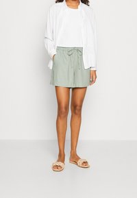 Shorts légers en lin vert avec taille ajustable à cordon, associés à un t-shirt blanc et une chemise blanche ample, complétés par des sandales beiges.