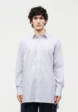 Άνδρας με σγουρά μαλλιά, ντυμένος με μεγάλη πουκαμίσα με λευκές και ανοιχτό μπλε ρίγες και σκούρο παντελόνι, στέκεται μπροστά σε απλό φόντο.