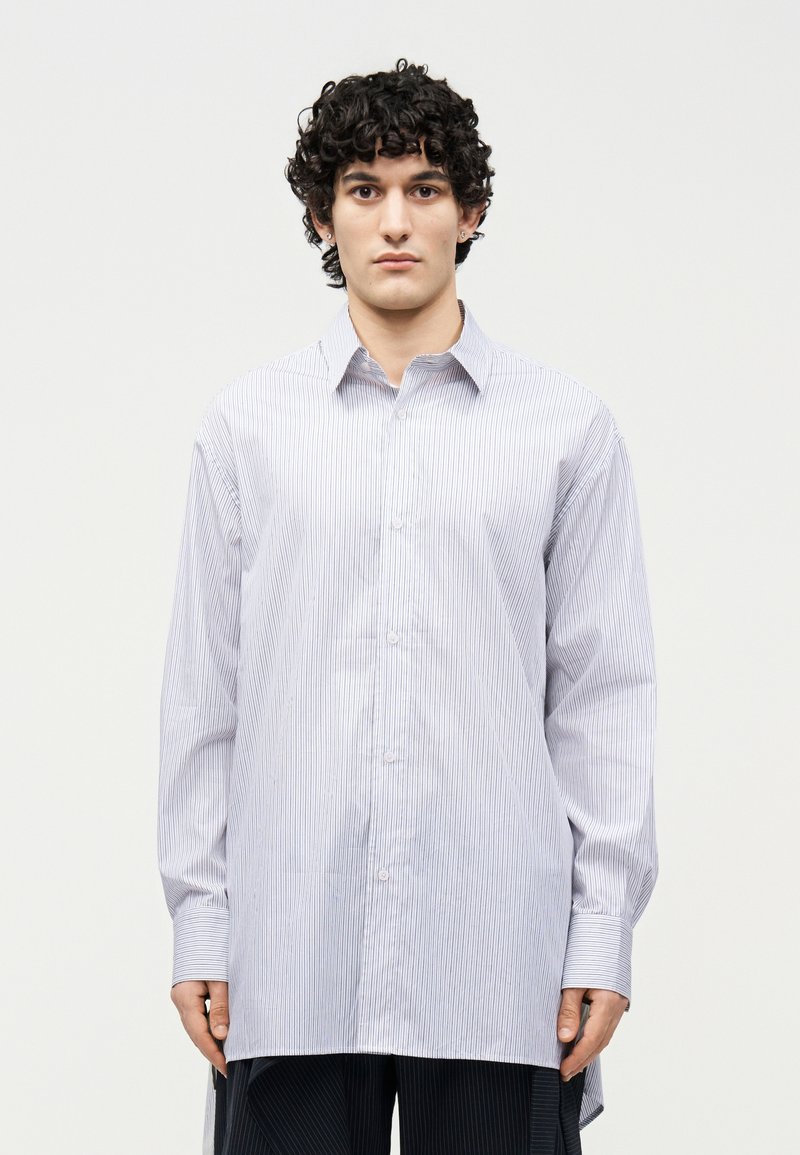 Άνδρας με σγουρά μαλλιά, ντυμένος με μεγάλη πουκαμίσα με λευκές και ανοιχτό μπλε ρίγες και σκούρο παντελόνι, στέκεται μπροστά σε απλό φόντο.