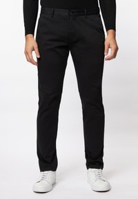 Zwarte slim-fit broek van een gladde stof, met een vlakke voorkant, riemlussen en een enkele knoopsluiting, gecombineerd met witte sneakers.