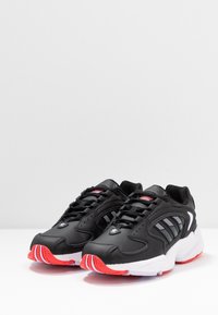 Black athletic shoes with multiple white stripes, red accents on the sole, and a textured leather upper. Features standard lace closure.