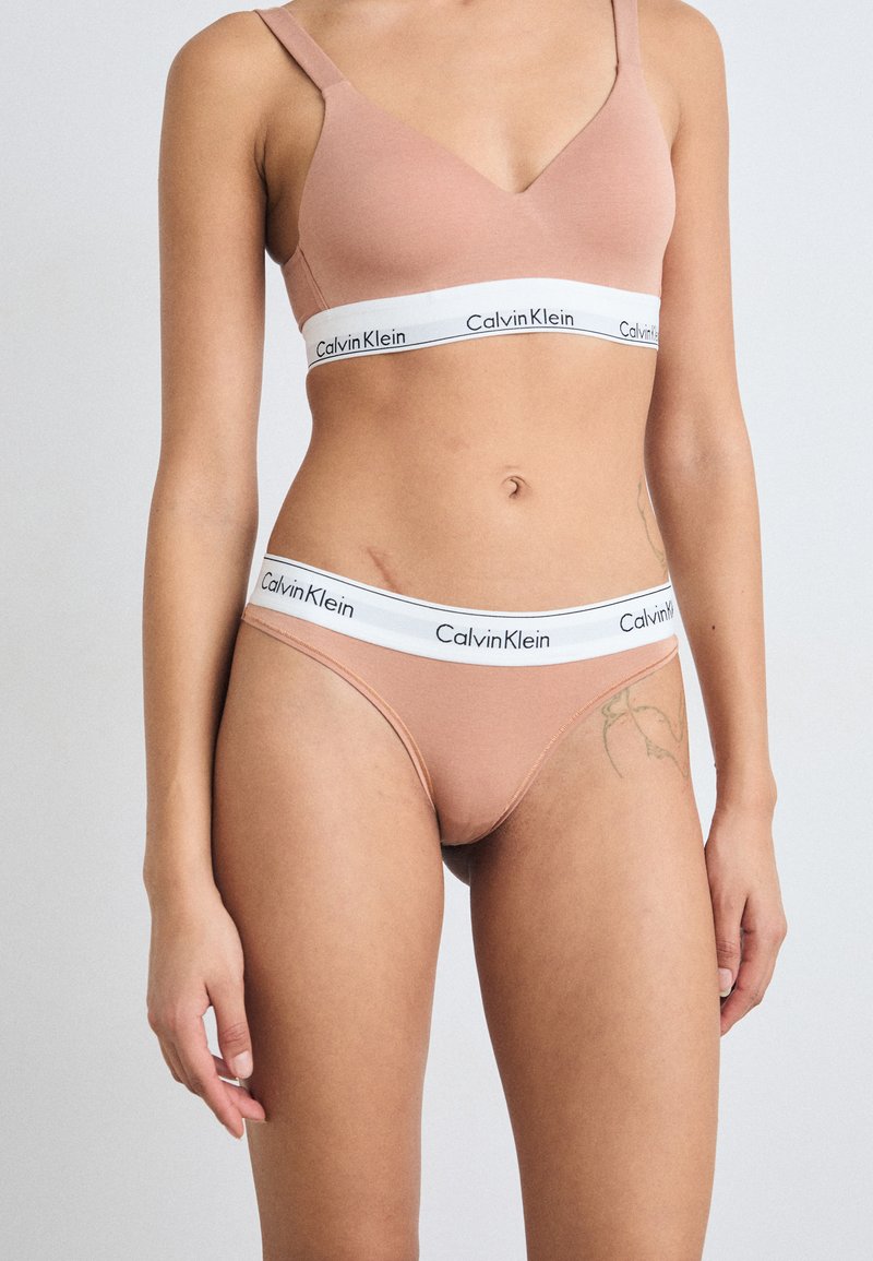 Calvin Klein bh en string set in zacht beige. Kenmerkt een v-hals ontwerp, dikke witte tailleband met logo, en glad, rekbaar materiaal.