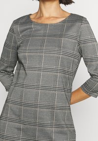 Černobílé kostkované šaty s tříčtvrtečními rukávy, kulatým výstřihem a texturovanou látkou s jemnými zlatými akcenty v motivu.