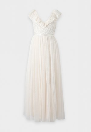 Mulher com cabelo longo e escuro a usar um vestido creme sem mangas com ombros franzidos e corpete de renda floral, combinado com sapatos creme com salto e tipo slip-on.