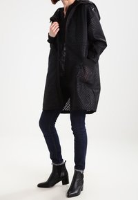 Manteau matelassé noir avec capuche, doublure en maille, poches latérales et coupe droite. Porté avec un jean foncé et des bottines à talons.
