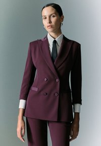 Giacca doppiopetto in tessuto bordeaux scuro, caratterizzata da un design su misura, revers a punta e polsini della camicia bianchi a contrasto.