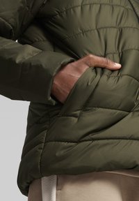 Olívazöld puffertakaró, vízszintes varrással, cipzáras elülső résszel és oldalsó zsebbel, sima, steppelt textúrával.