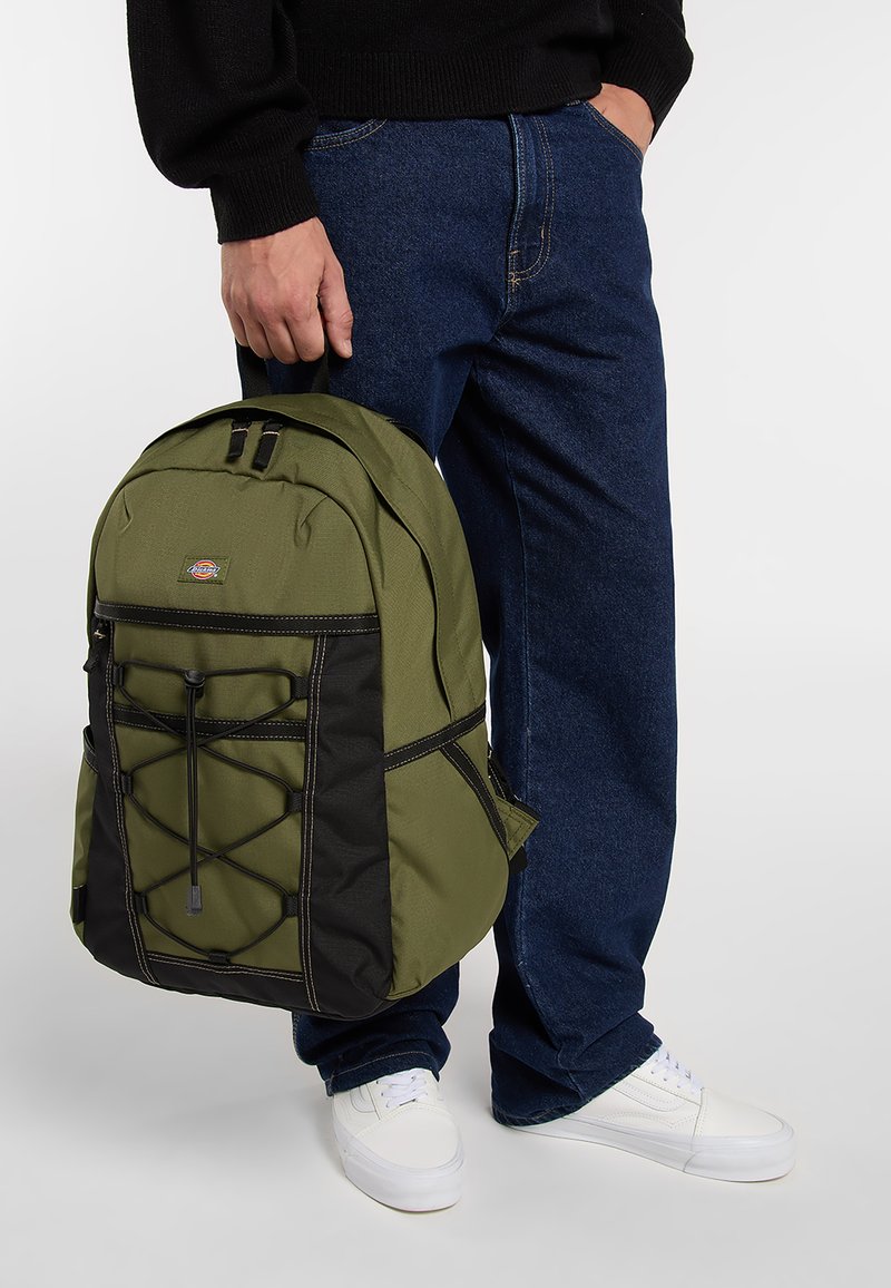 Mochila verde oliva con detalles en negro, que presenta una tela texturizada, dos asas superiores y una cuerda elástica en la parte frontal.