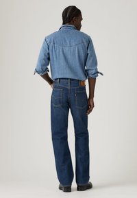 Ανοιχτό μπλε πουκάμισο τζιν με μανσέτες με κουμπιά, φορεμένο πάνω από σκούρο μπλε τζιν παντελόνι. Ορατές πίσω τσέπες, ελαφρώς φαρδιά εφαρμογή και μαύρα μποτάκια.