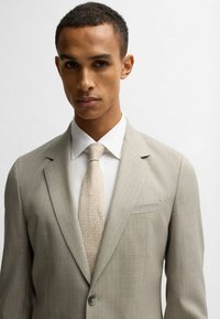 Completo grigio chiaro con cravatta beige texture, camicia bianca e chiusura a un bottone. I dettagli notevoli includono sottili righe verticali e revers.