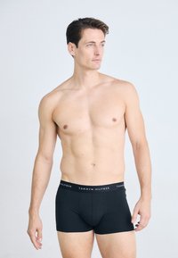 Shirtless man with short brown hair wearing black Tommy Hilfiger boxer briefs, standing against a plain light background, looking right.