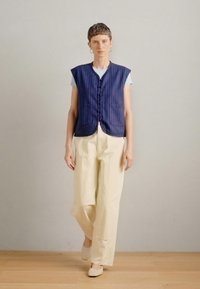 Tamsiai mėlynos ir baltos juostelės be rankovių liemenė su sagomis, užvilkta ant šviesiai mėlynos marškinėlių ir derinama su dramatiškai baltais plačiais kelnais ir šviesiai rudos spalvos batais.