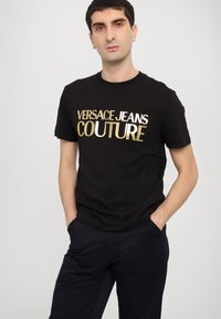 Juodas medvilninis marškinėliai su dideliu auksiniu metalo spausdinimu, ant kurio užrašyta "VERSACE JEANS COUTURE." Trumpomis rankovėmis, suapvalinta kaklo iškirpte, laisvas kirpimas.