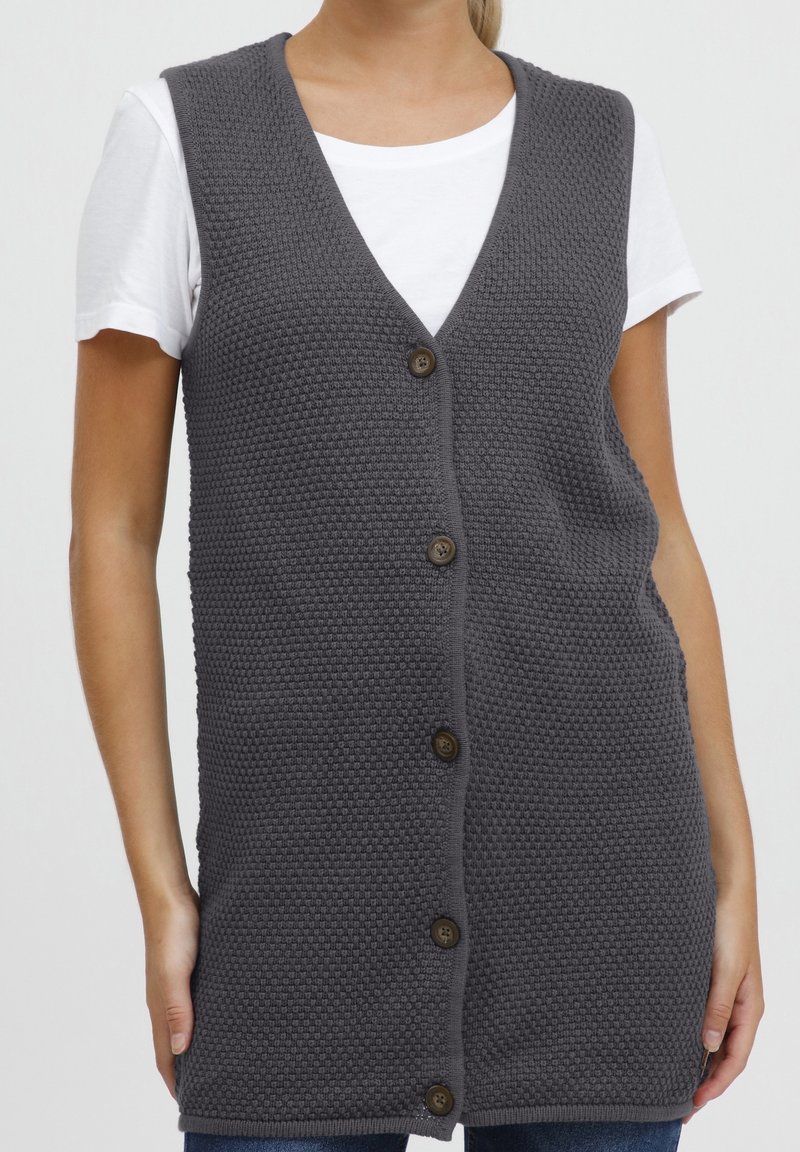 Graue, gestrickte Weste mit strukturierter Oberfläche, V-Ausschnitt und fünf braunen Knöpfen vorne. Wird über einem weißen, ärmellosen Hemd getragen.