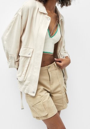 Vrouw draagt een beige oversized jas, een gebreid cropped top met groene rand en beige cargoshorts tegen een witte achtergrond.