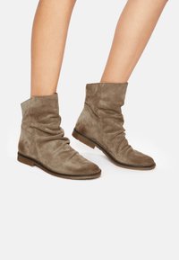 Bottines en daim marron avec un design froncé, semelle plate et bout arrondi, présentant une finition texturée et des détails minimalistes.