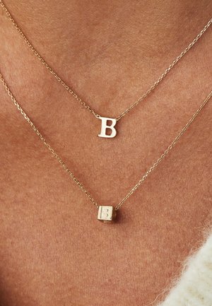 Colliers en couches de couleur or avec un pendentif circulaire lisse portant un "B" et un pendentif en forme de cube gravé d'un "B", mettant en valeur des chaînes délicates.
