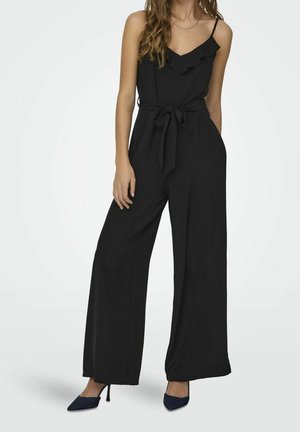 Femme portant une combinaison noire sans manches avec des jambes larges, une ceinture nouée à la taille et des chaussures à talons pointus bleu marine, debout sur un fond uni.