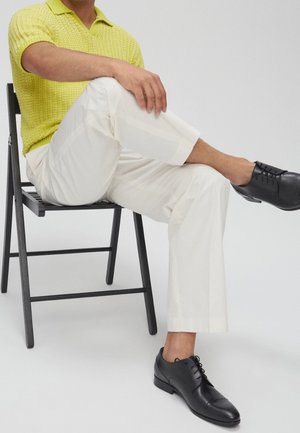Chaussures en cuir noir, pantalon blanc et polo jaune texturé ; posture assise sur une chaise noire, fond neutre.