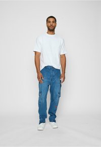 Weißes kurzärmliges T-Shirt kombiniert mit blauen Cargo-Jeans, die zwei Seitentaschen und ein kleines rotes Logo aufweisen. Die Schuhe sind weiße Sneaker.