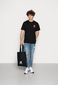 Svart T-skjorte med rødt sirkulært design og tekst, slitte lyseblå jeans, hvite sneakers med røde og blå detaljer, svart totebag.