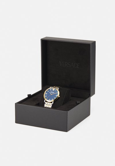 Hodinky Versace v čiernej krabičke; zlatý rám, modrý ciferník so striebornými akcentmi, nerezový náramok, umiestnené na čiernom zamatovom vankúši.