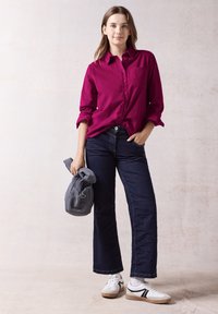 Magenta button-up shirt with a chest pocket, paired with dark blue wide-leg jeans and white sneakers with black accents. Holding a gray bag.