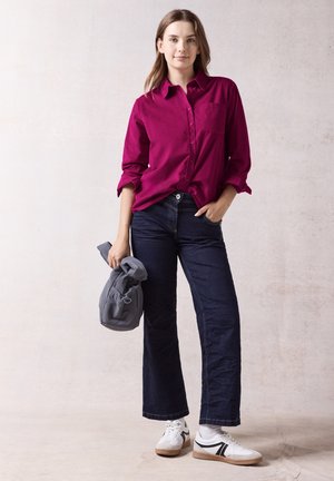 Magenta-Knopfhemd mit einer Brusttasche, kombiniert mit dunkelblauen weiten Jeans und weißen Sneakers mit schwarzen Akzenten. Hält eine graue Tasche.