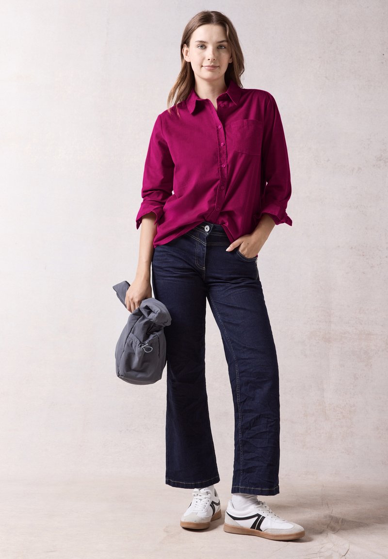 Magenta button-up shirt with a chest pocket, paired with dark blue wide-leg jeans and white sneakers with black accents. Holding a gray bag.