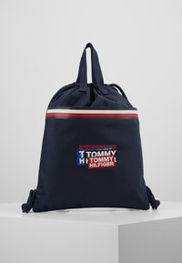 Bolsa de cordón azul marino con doble asa, rayas rojas, blancas y azules cerca de la parte superior, y parches del logo de Tommy Hilfiger en capas en la parte frontal.