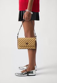 Borsa a strisce oro e bianca con logo triangolare, rifiniture marroni e una tracolla rimovibile. Un bracciale trasparente è indossato al polso.