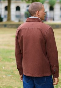 Brun bomuldsjakke med krave og trykknapper, med en afslappet pasform og rygstykke. Sammen med blå denim jeans, udendørs.