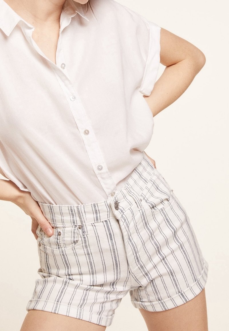 Personne portant une chemise blanche à manches courtes boutonnée et un short blanc taille haute avec des rayures verticales grises, les mains sur les hanches.
