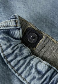 Šviesiai mėlynos denimo medžiagos paviršius turi siuvinėtą kraštą, tekstūruotą etiketę ir tamsų mygtuką su keturių skylių dizainu.