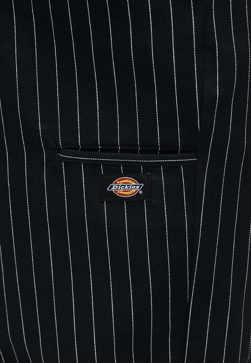 Schwarzer Stoff mit dünnen weißen vertikalen Nadelstreifen, einer kleinen horizontalen Schlitztasche und einem rechteckigen Dickies-Markenlabel unterhalb der Tasche.