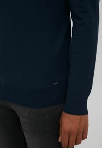 Suéter de punto azul marino con puños y dobladillo acanalados, que presenta una pequeña etiqueta de metal en la parte inferior izquierda. Lleva puesto sobre unos jeans negros.