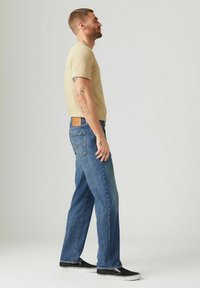 Camiseta de algodón beige de manga corta, combinada con jeans de denim azul de pierna recta, con un parche de cuero en la espalda, llevada con zapatos negros.