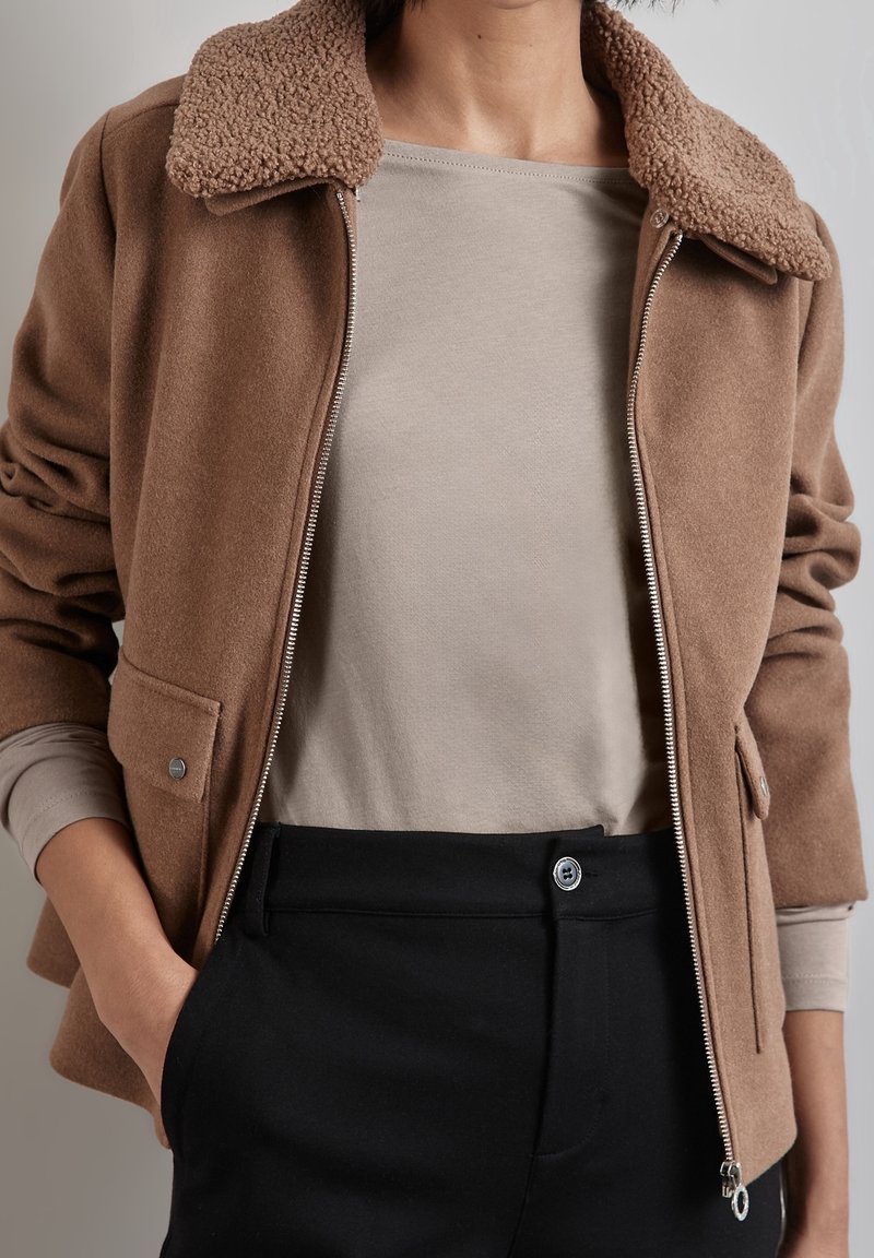 Brun ullblandad jacka med en strukturerad beige krage, dragkedja framtill och sidofickor, lager på lager över en ljusbeige topp och svarta byxor.