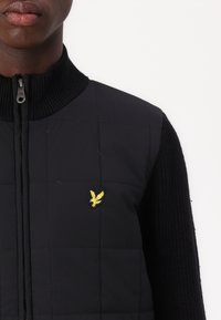 Black quilted vest with ribbed collar and sleeves. Features yellow embroidered eagle logo on the upper left chest. Zipper closure.