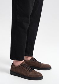 Baskets en daim marron avec des lacets noirs, présentant un profil bas et une semelle en caoutchouc. Portées avec un pantalon noir ajusté pour un look décontracté.