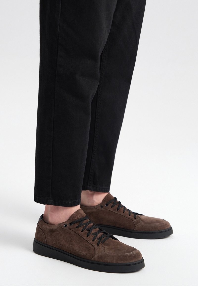 Baskets en daim marron avec des lacets noirs, présentant un profil bas et une semelle en caoutchouc. Portées avec un pantalon noir ajusté pour un look décontracté.