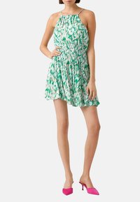 Robe halter à motifs verts et blancs avec une taille smockée, longueur au-dessus du genou et fines bretelles. Associée à des talons roses vifs.